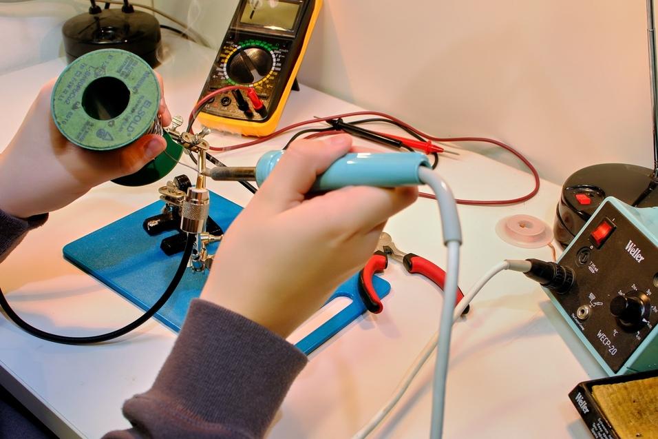 es sind Hände zu sehen, ein Messgerät, eine "Dritte-Hand" in die ein Koaxkabel mit Stecker eingeklemmt ist., die rechte Hand hält einen Lötkolben und lötet einen Stecker an. es sind Hände zu sehen, ein Messgerät, eine "Dritte-Hand" in die ein Koaxkabel mit Stecker eingeklemmt ist., die rechte Hand hält einen Lötkolben und lötet einen Stecker an.