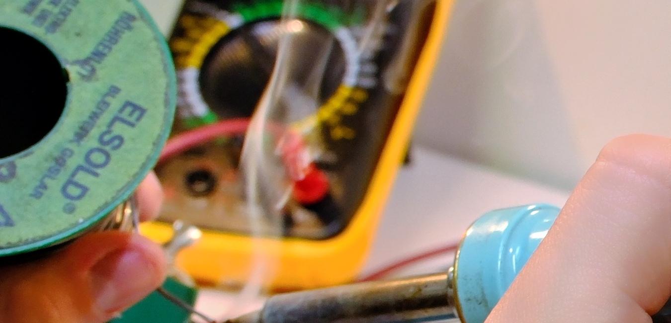 im Hintergrund ein Messgerät, im Vordergrund hält die rechte Hand einen Lötkolben, es wird ein PL-Stecker gelötet, Lötdampf steigt auf!