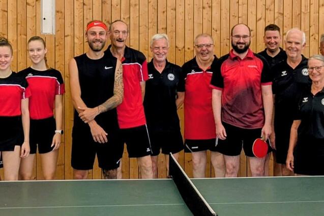 Fünfzehn Tischtennisspielerinnen und -spieler in roter und schwarzer Vereinskleidung stehen in einer Sporthalle hinter einem grünen Tischtennistisch. Die Gruppe umfasst Männer und Frauen verschiedener Altersgruppen.