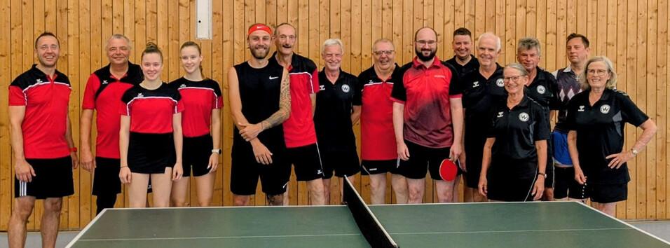Vor den Freundschaftsspielen - SVW und TTC Großdietmanns Fünfzehn Tischtennisspielerinnen und -spieler in roter und schwarzer Vereinskleidung stehen in einer Sporthalle hinter einem grünen Tischtennistisch. Die Gruppe umfasst Männer und Frauen verschiedener Altersgruppen.