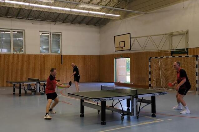 Spieler in Aktion an der Tischtennisplatte in der Halle: Ein Spieler im roten Trikot schl&auml;gt einen Vorhand-Return, w&auml;hrend sein Gegner am gegen&uuml;berliegenden Ende der Platte returniert bereitsteht. Im Hintergrund sind zwei weitere Spieler an