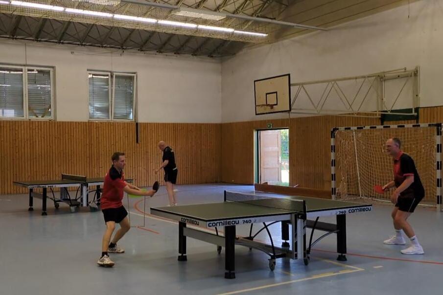 Spieler in Aktion an der Tischtennisplatte in der Halle: Ein Spieler im roten Trikot schlägt einen Vorhand-Return, während sein Gegner am gegenüberliegenden Ende der Platte returniert bereitsteht. Im Hintergrund sind zwei weitere Spieler an
