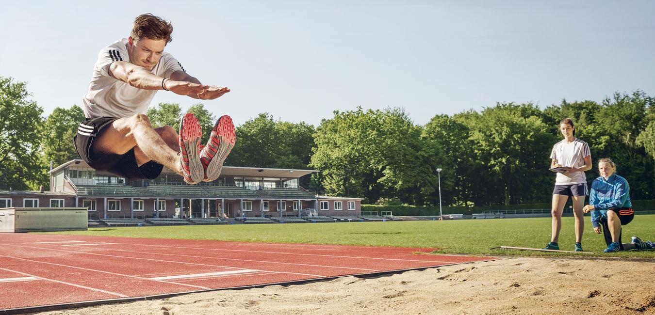 Weitspringer in der Luft beim Absprung über eine Sandgrube, daneben zwei Beobachterinnen.