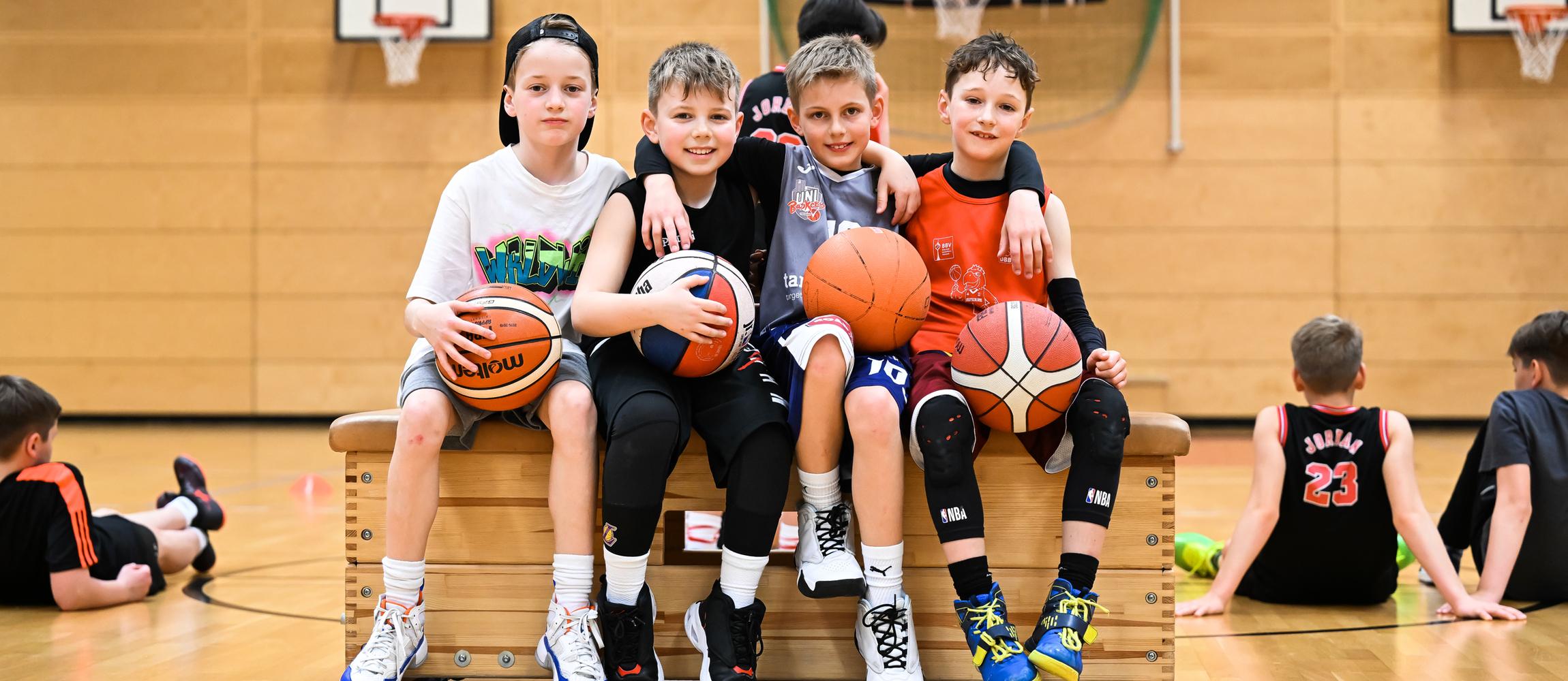 Vier Kinder sitzen auf einer Bank in der Turnhalle, halten Basketballs und lachen. Im Hintergrund sind weitere Kinder aktiv.