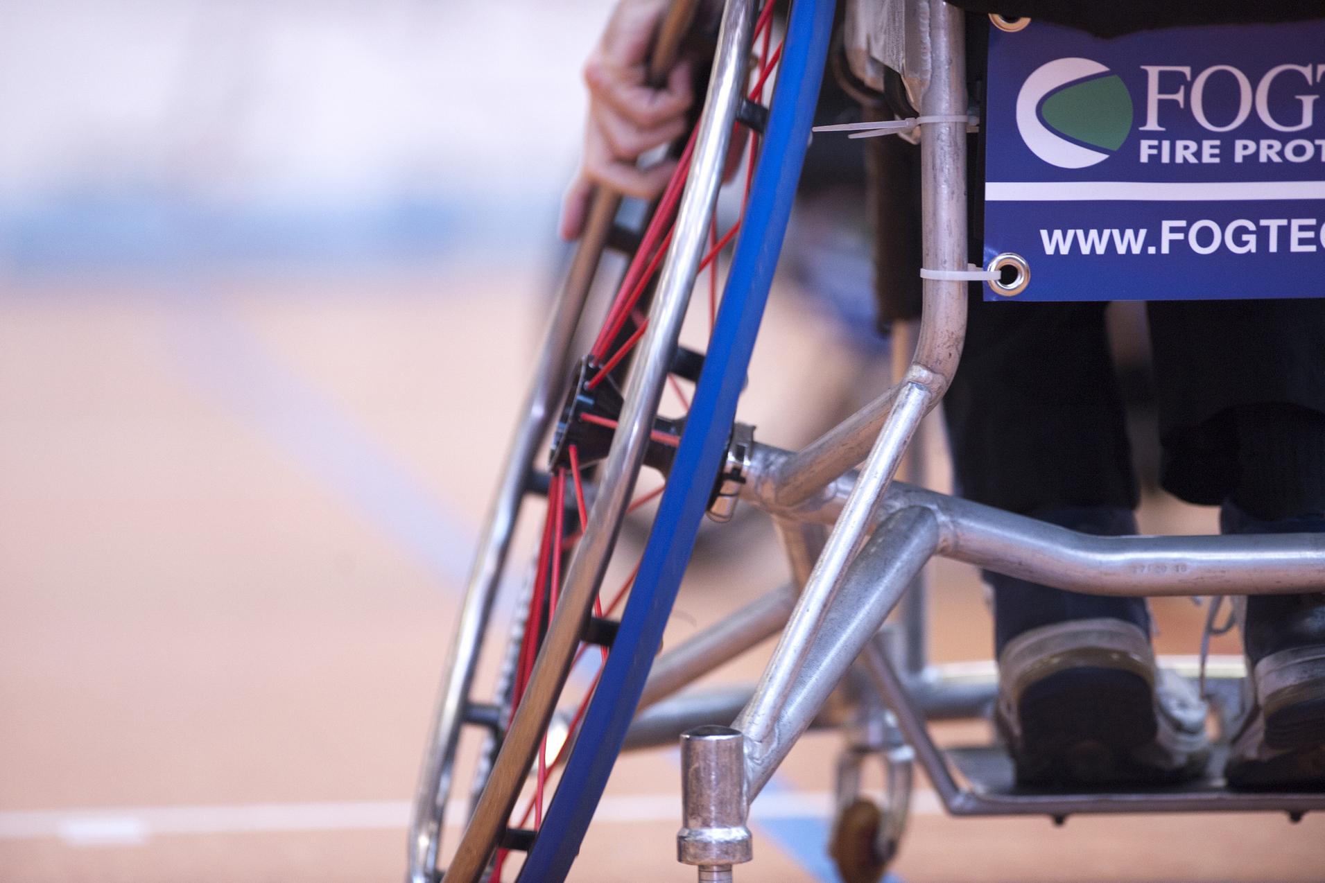 Detailansicht eines Rollstuhls mit einem leuchtend blauen und roten Rad sowie einem Werbeschild auf der Rückenlehne.