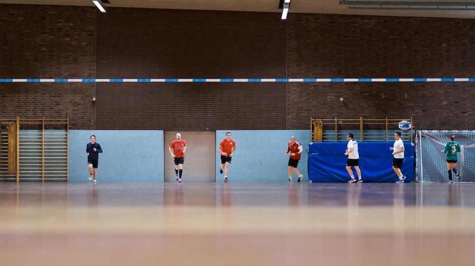Fünf Spieler in Sportkleidung trainieren in einer Sporthalle, mit einem Tor und blauen Wänden im Hintergrund.