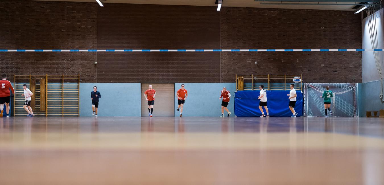 Fünf Spieler in Sportkleidung trainieren in einer Sporthalle, mit einem Tor und blauen Wänden im Hintergrund.