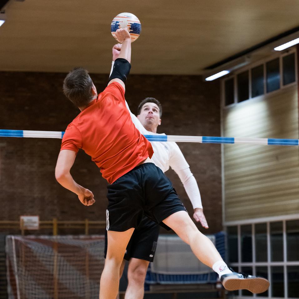 Zwei Spieler im Sprung während eines Handballspiels, einer wirft den Ball über eine Markierung.