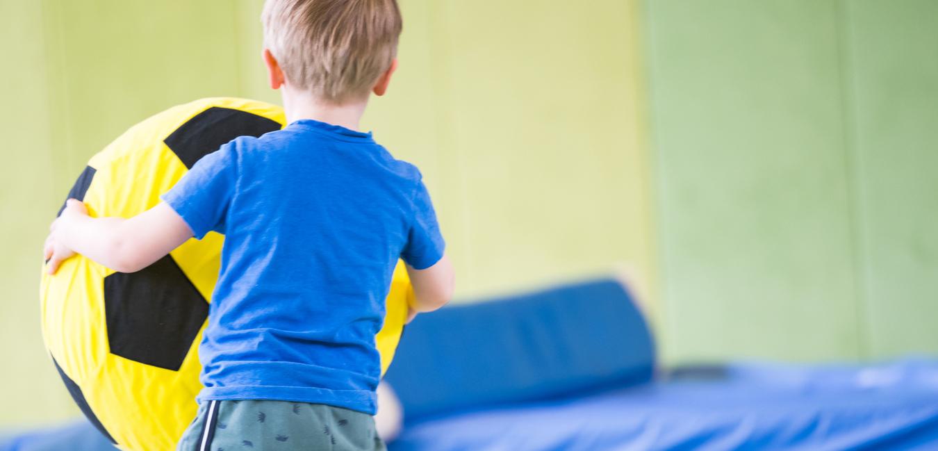 Ein Junge trägt einen großen, gelben Ball mit schwarzen Feldern in einer Turnhalle.