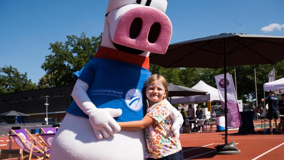 Ein Kind umarmt ein Maskottchen in Sportbekleidung auf einem Sportplatz mit Ständen und Sportequipment im Hintergrund.