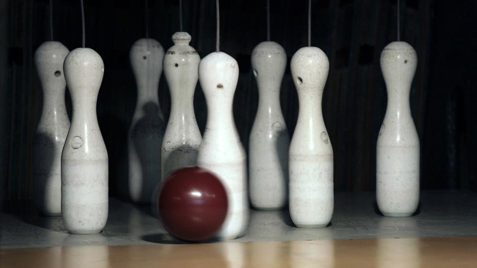 Eine rotbraune Bowlingkugel rollt auf eine Reihe weißer Kegel auf einer Bowlingbahn zu.