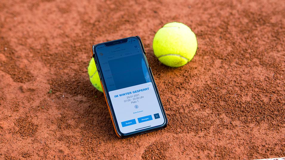 Smartphone mit Buchungsinfo liegt auf einem Tennisplatz, neben zwei gelben Tennisbällen auf rotem Sand.
