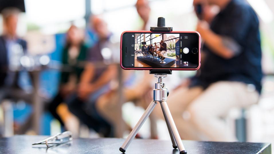 Smartphone auf einem Stativ, das eine Gruppe von Personen in einem modernen Raum aufnimmt. Unscharfer Hintergrund.