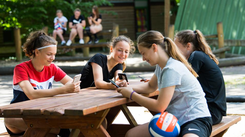 Vier Mädchen sitzen an einem Holztisch und nutzen ihre Smartphones, während im Hintergrund andere Kinder spielen.