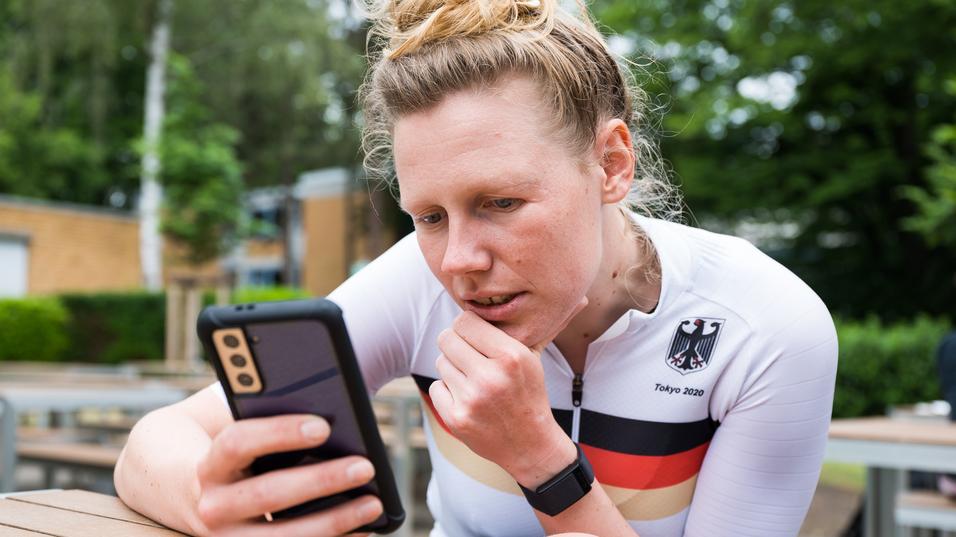 Athletin im weißen Trikot mit schwarzem und rotem Streifen schaut konzentriert auf ihr Smartphone in einer Außenanlage.