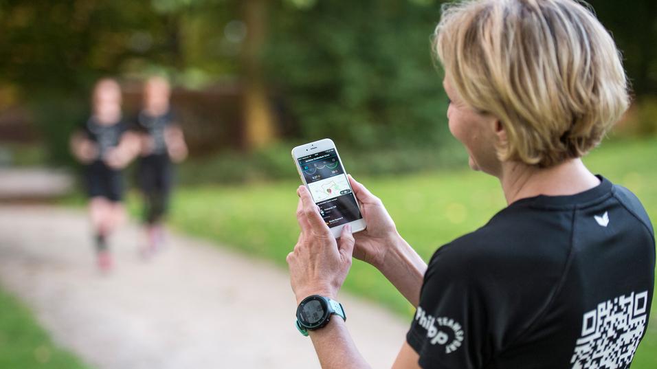 Frau mit kurzem Haar schaut auf ihr Smartphone, während zwei Jogger im Hintergrund auf einem Weg vorbeilaufen.