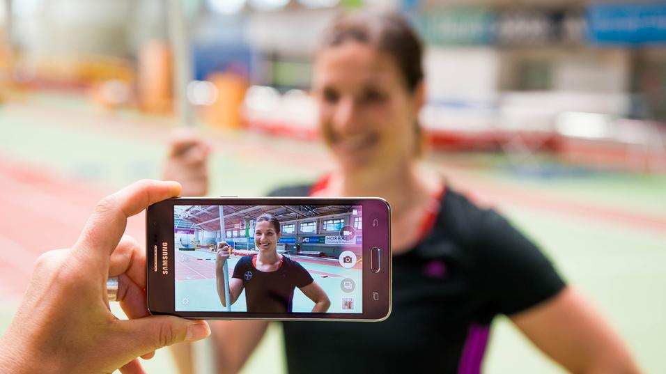 Hand hält ein Smartphone, das ein Porträt einer lächelnden Athletin mit Stab in einer Sporthalle zeigt.