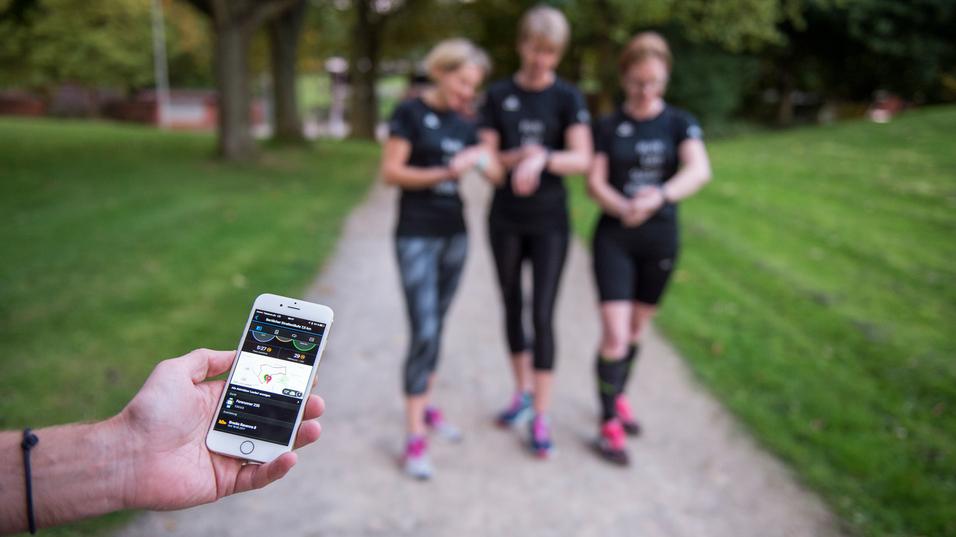 Ein Mann hält ein Smartphone mit Fitness-App, während drei Frauen in Sportkleidung auf einem Spazierweg stehen.