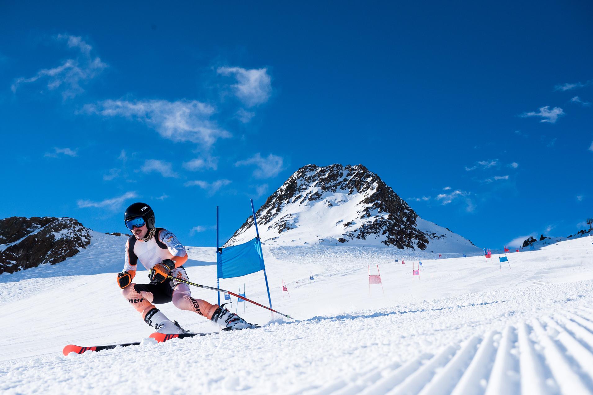 Ski fahrender Athlet auf einer verschneiten Piste mit Toren und einem Berg im Hintergrund unter blauem Himmel.