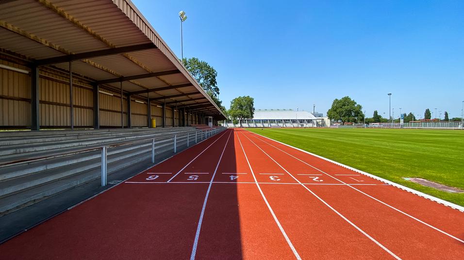 Leere Laufbahn mit roten Bahnen und Tribüne unter einem klaren blauen Himmel, neben einem Kunstrasenfeld.