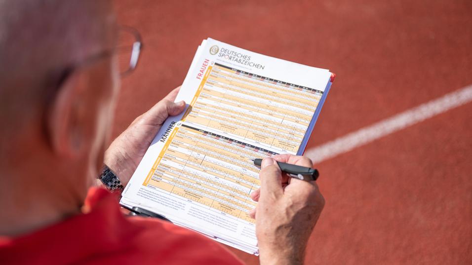 Hand eines Prüfers, der auf einem Blatt mit Sportabzeichen-Anforderungen auf einer Laufbahn notiert.