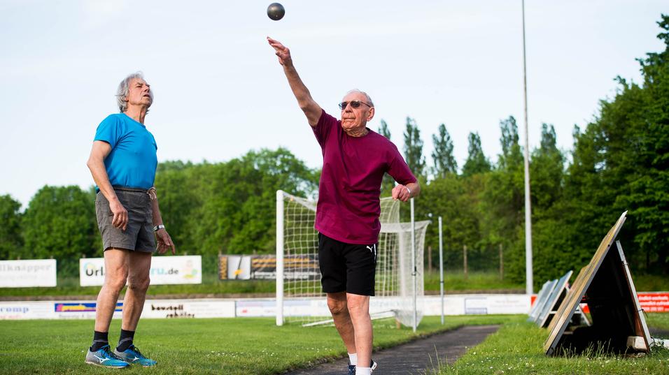 Zwei ältere Männer üben das Kugelstoßen auf einem Sportplatz, einer wirft die Kugel, der andere beobachtet.