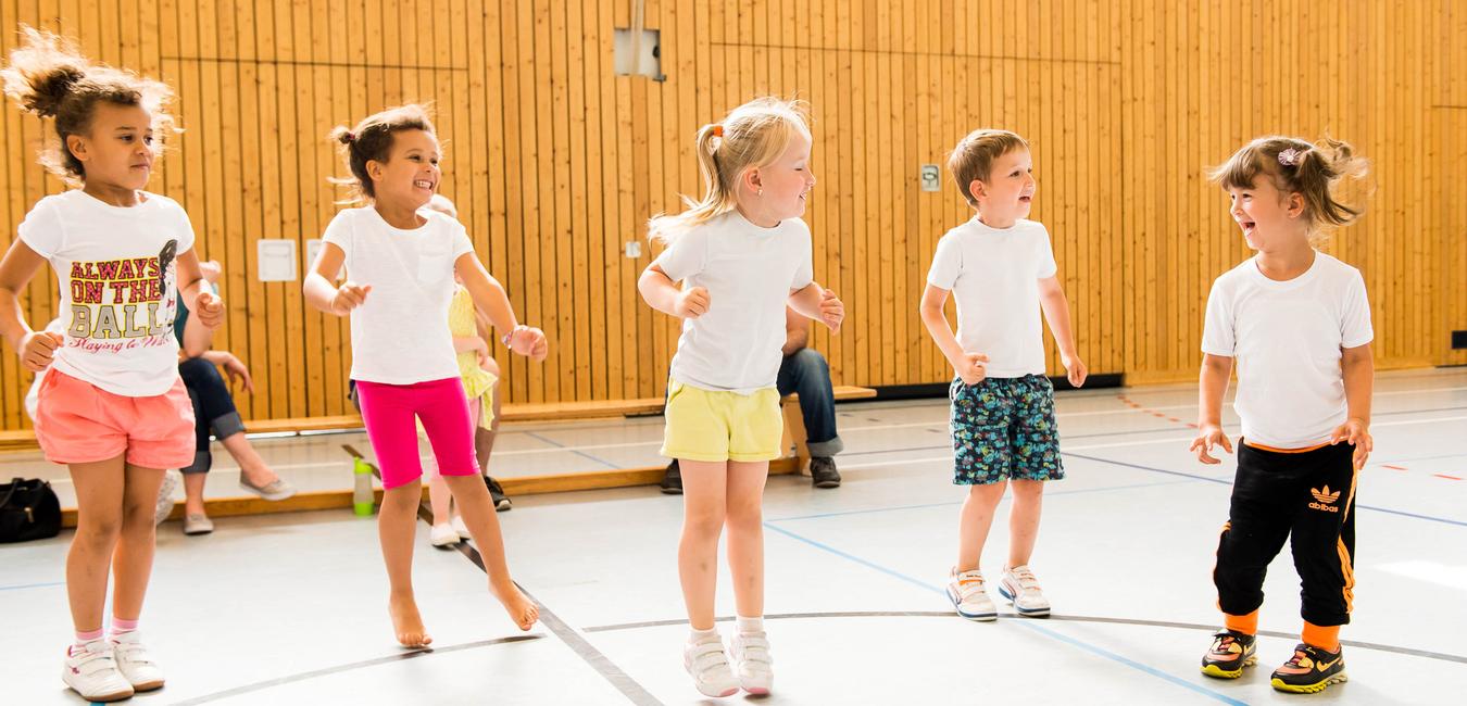 Fünf Kinder in T-Shirts springen fröhlich in einer Sporthalle mit Holzwänden und einen Zuschauer im Hintergrund.