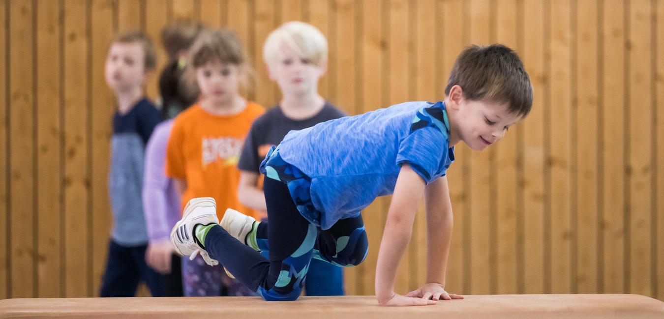 Ein Junge balanciert auf einem Blickkasten, während mehrere Kinder im Hintergrund zuschauen. Holzpaneele sind sichtbar.
