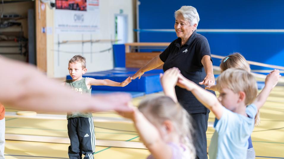 Seniorin leitet Kinder in einer Sporthalle an, während sie ihre Arme in die Höhe strecken.