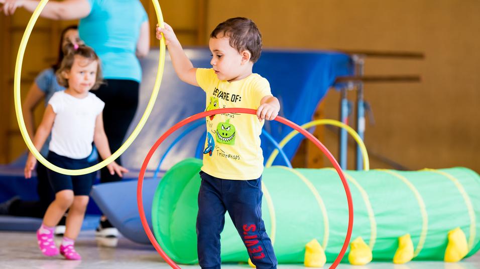 Zwei Kinder spielen in einer Turnhalle mit hula-hoops und einem grünen Tunnel. Ein Kind hält zwei Reifen in der Hand.