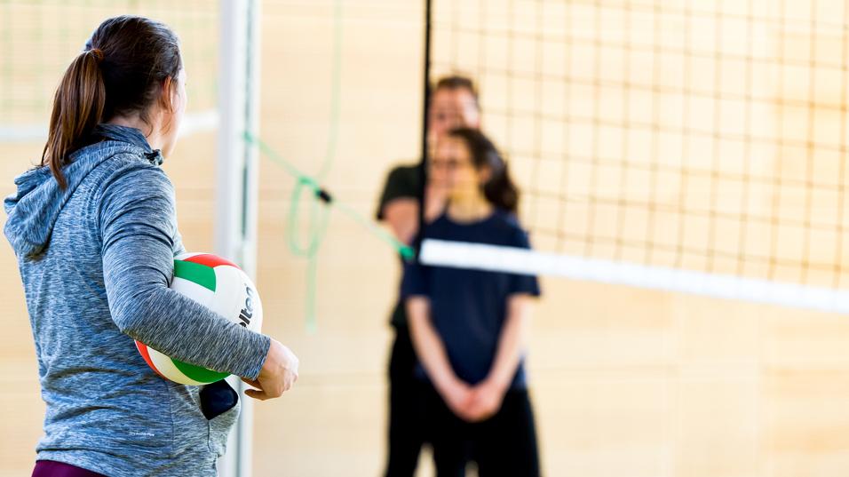 Eine Person mit einem Volleyball steht vor dem Netz, während zwei weitere Personen im Hintergrund warten.