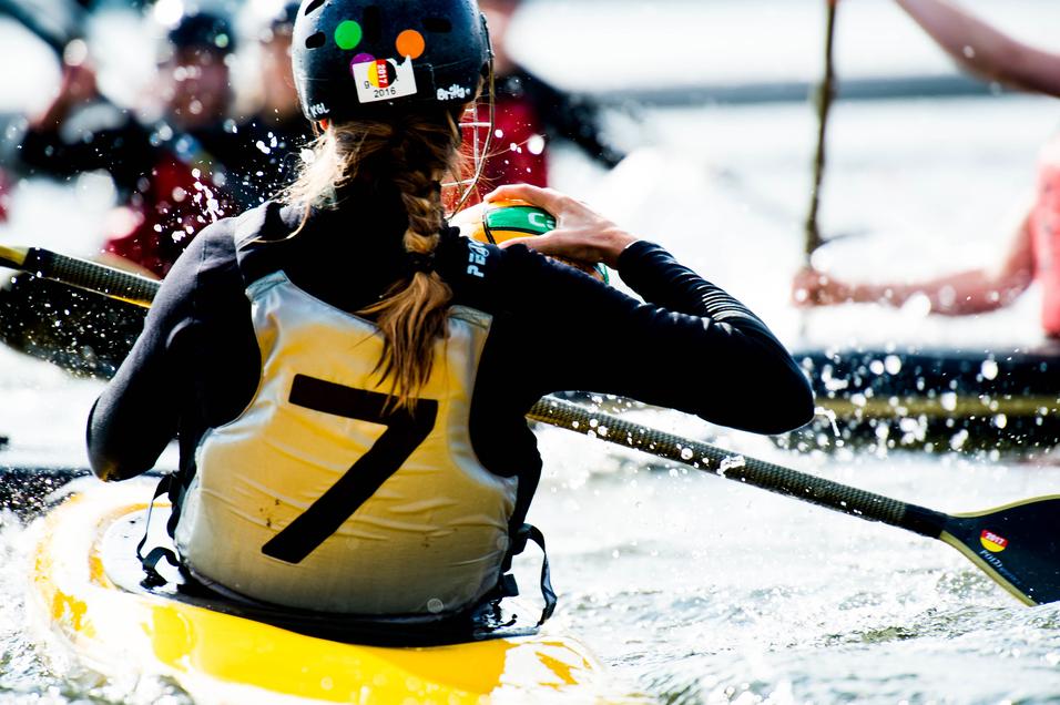Nahaufnahme einer Paddlerin in einem gelben Kajak, die einen Ball hält, während Wasser spritzt.