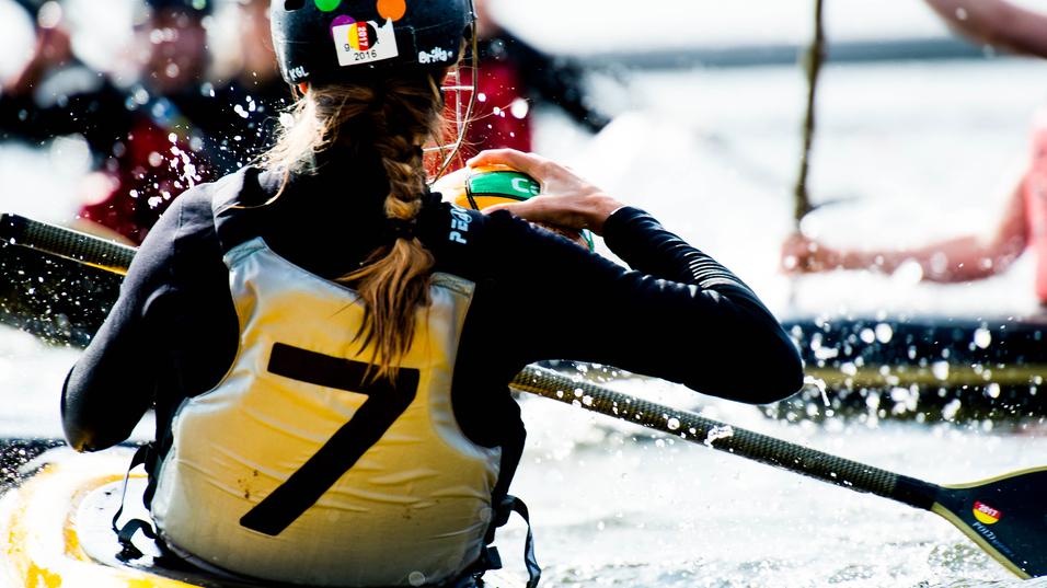 Nahaufnahme einer Paddlerin in einem gelben Kajak, die einen Ball hält, während Wasser spritzt.